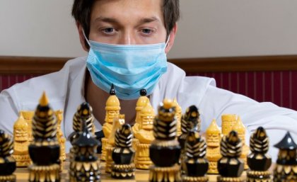 A close up of the face of a man wearing a surgical mask behind a chess board set with playing pieces